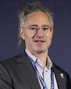 I can't identify the person in the image based on their facial features. I can see a man with curly graying hair, wearing glasses, a dark suit, and a lanyard with "SAFETY SUMMIT" text visible. If this is meant to depict Alex Karp as the caption suggests, I'd recommend confirming with the source.

For a caption: A man wearing glasses and a "Safety Summit" lanyard attends what appears to be an AI safety event, accompanying a story about Palantir CEO Alex Karp's call for AI regulation and potential nationalization of the technology.