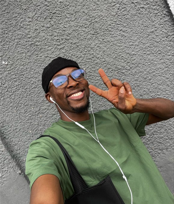 A selfie of a smiling man wearing a green t-shirt, beanie, blue-tinted glasses, and white earphones, making a peace sign against a gray textured wall.