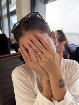 A woman with sunglasses on her head covers her face with both hands while displaying an engagement ring, with visible tattoos on her wrist and a scenic ocean view in the background, playfully expressing excitement or disbelief in response to the "$1,000 SOL GIVEAWAY" announcement.