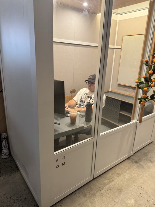 A man wearing a cap and glasses works at a desk inside a glass-walled booth labeled 'ROOM'.