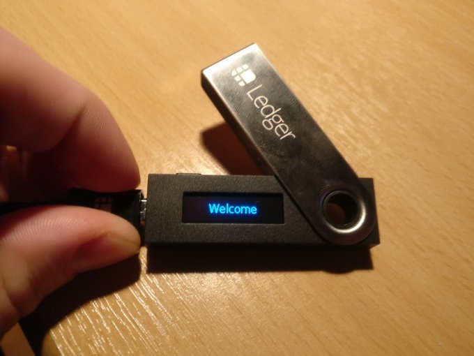A hand holds a USB cable plugged into a Ledger hardware wallet displaying the word 'Welcome' in bright blue text on its small screen, resting on a wooden surface.