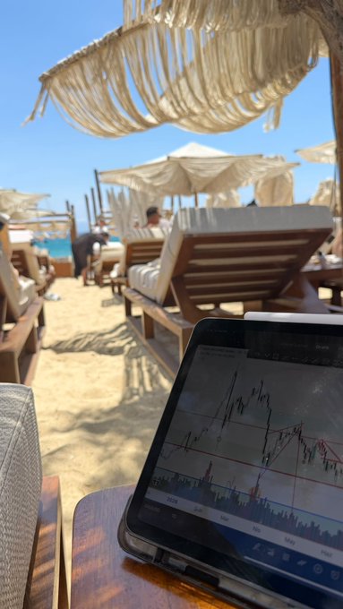 The image shows a laptop computer sitting next to a beach chair with an umbrella and a surfboard hanging off the back of the chair. The scene has elements of both relaxation and productivity, as people may be enjoying their time at the beach while also using technology like laptops for work or communication purposes.