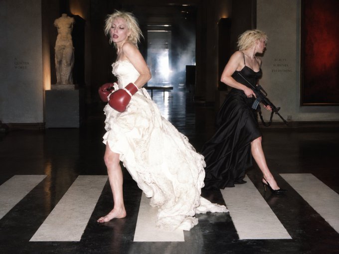A dramatic fashion editorial photo shows two women in formal dresses—one in white with red boxing gloves, the other in black holding a rifle—striking action poses on a crosswalk in a museum-like hall.