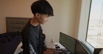 A young man sits at a computer setup with dual monitors in a modern apartment by a large window, appearing focused on his screen. The image likely depicts the college student mentioned in the post, who became an internet sensation for allegedly profiting $40 million from $TRUMP trading.