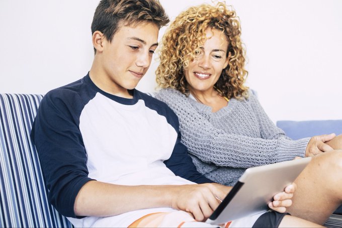 A smiling woman with curly blonde hair and a teenage boy sit together on a striped couch, looking down at a tablet the boy is holding.