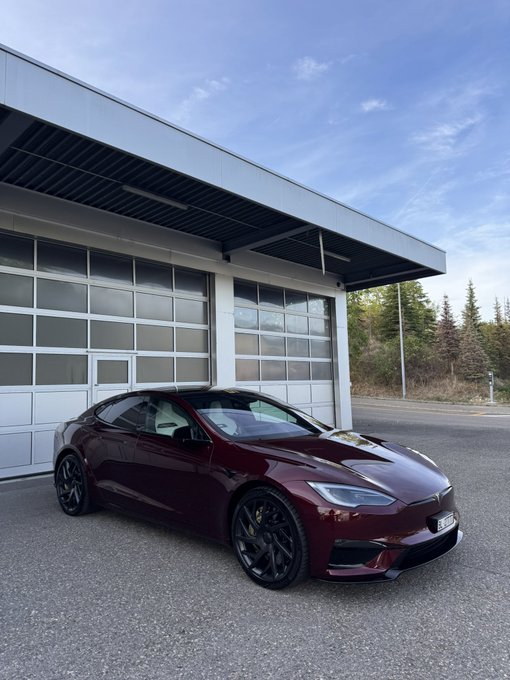 A dark maroon Tesla Model S parked in front of a building with large glass garage doors under a blue sky.