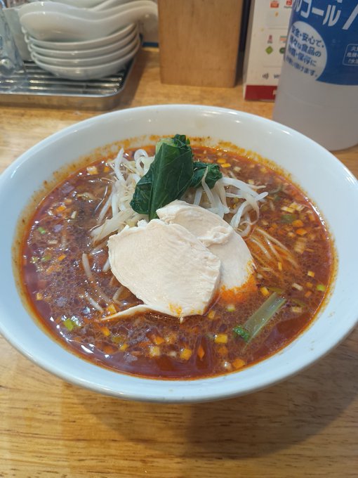 A bowl of spicy red noodle soup topped with sliced chicken, bean sprouts, and greens sits on a wooden table next to a bottle of Japanese beverage.