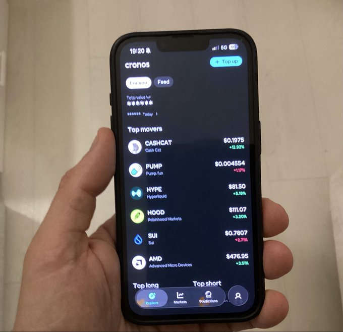 A hand holds a smartphone displaying the Crypto.com app with the 'Top movers' list, showing gains for tokens like CASHCAT (+13.92%) and HOOD (+3.20%), and a drop for AMD (-3.51%).