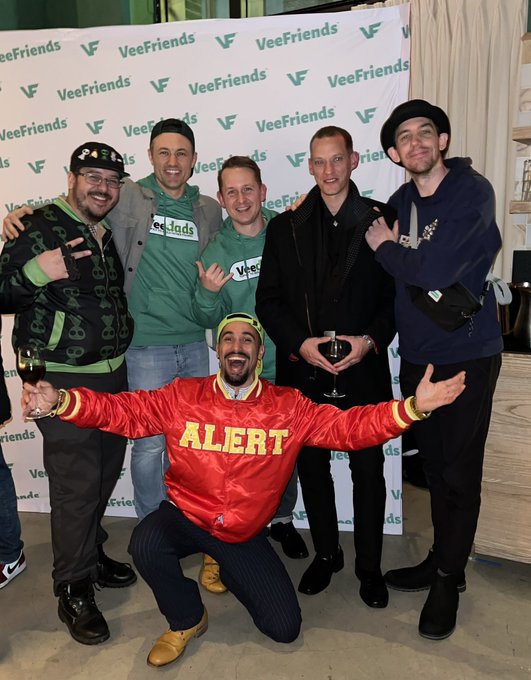 A group of six men posing in front of a VeeFriends-branded backdrop, with one man kneeling in front wearing a red satin 'ALERT' jacket and yellow cap, while others wear green VeeFriends apparel and hold drinks.