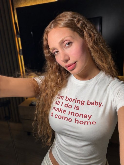 A young woman with long curly blonde hair takes a selfie wearing a white t-shirt that reads 'i'm boring baby, all I do is make money & come home' in red text.