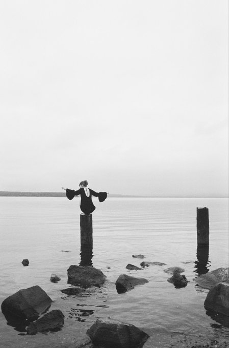 A black-and-white photograph shows a figure in dark clothing standing with arms outstretched atop a wooden post in calm water, with rocks scattered in the foreground and another post to the right.