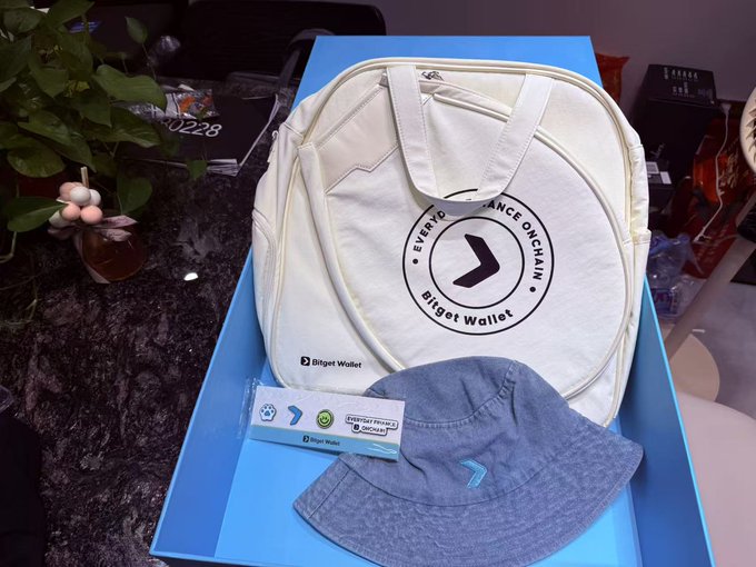 A Bitget Wallet promotional package containing a cream-colored duffel bag with the 'EVERY DANCE ON CHAIN' logo, a gray bucket hat, and stickers, displayed on a blue box.