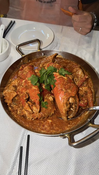 A large metal pan filled with crab in a rich reddish-orange sauce garnished with cilantro, served on a white tablecloth with chopsticks and empty plates nearby.
