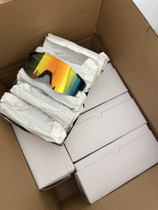 An open cardboard shipping box containing white product packaging and sunglasses with a rainbow/iridescent reflective lens.