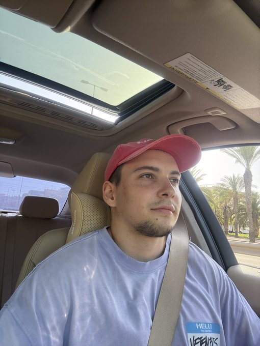 A selfie of a young man wearing a red cap and light blue sweatshirt sitting in the passenger seat of a car with a sunroof open, palm trees visible through the window.