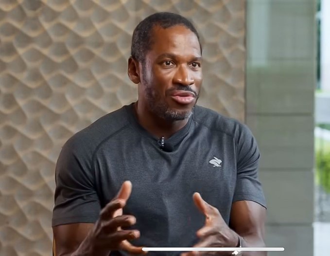 The image features an interview with a man wearing a gray shirt sitting at a table. He has his hands outstretched as he speaks, giving the impression that he is engaged in conversation or sharing information. The background of the image shows a wall with a unique texture resembling waves, adding to the overall atmosphere of the scene.
