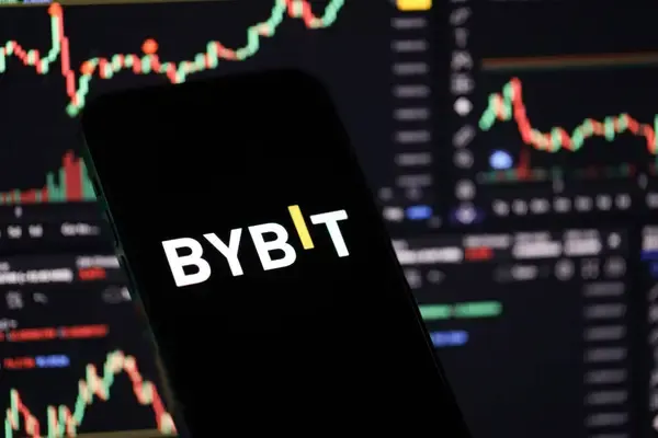 The Bybit logo displayed on a smartphone screen in front of trading charts visually reinforces the article's message about the cryptocurrency exchange expanding into comprehensive banking services beyond just crypto trading under Europe's MiCA regulations.