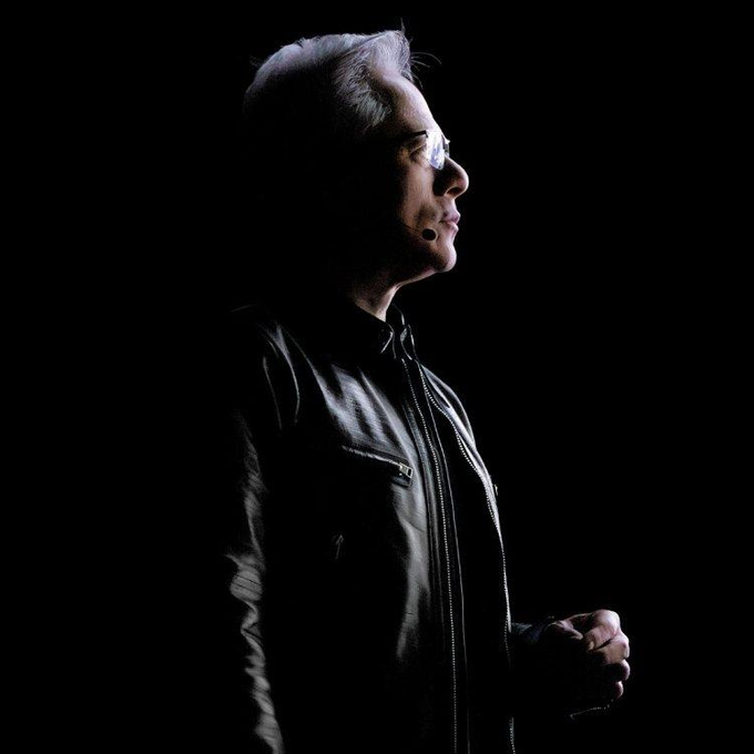 A man with silver hair and glasses stands in profile against a black background, illuminated by dramatic side lighting that highlights his iconic black leather jacket. The image embodies the post's message—while executives swap tailored suits to fit trends, true cultural icons like him remain instantly recognizable in their signature look, a symbol of timeless identity rather than conformity.