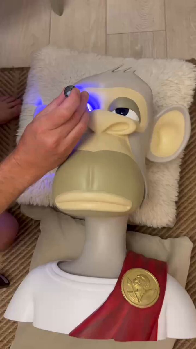 The image features a man's hand holding up a toy monkey with blue eyes, which appears to have been made from resin or plastic. The monkey has a red and gold robe around its neck, giving it an authentic appearance. The caption of the image reads "Gm gm Happy Mutant Monday Here is a @BoredApeYC bust getting his eyeballs made out of resin He will be finished soon and ready to fly to his new home!!".