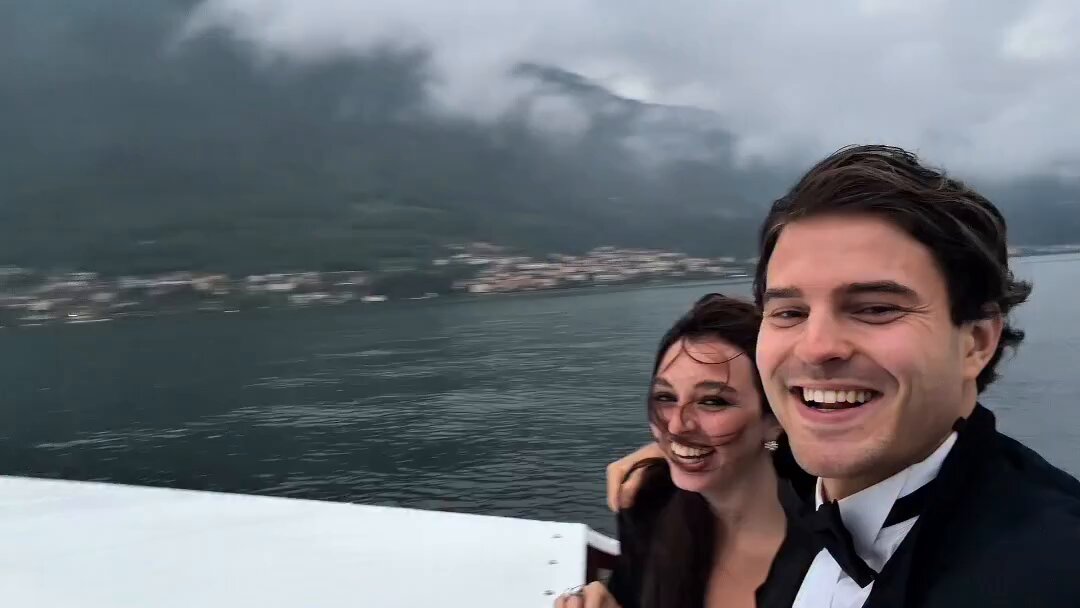 The image shows a man wearing a tuxedo and a woman dressed in black, both smiling at the camera while standing on a boat with their backs to the viewer. The background features a cloudy sky over a body of water, possibly an ocean or lake. There is also text visible in the image that reads "I’m living off my entire bags of $XRP".