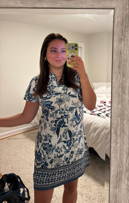 A woman in a blue and white floral dress takes a mirror selfie with a yellow iPhone in a bedroom, smiling as she greets "crypto twitter" with a cheerful "good morning" and a heart. The casual, fresh-outfit vibe mirrors the upbeat, friendly tone of the morning post to her followers.