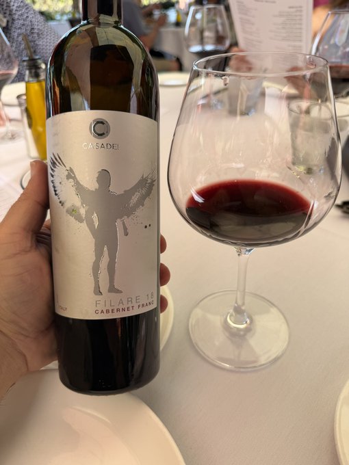 A hand holding a bottle of 'CASADEI FILARE 18 CABERNET FRANC' red wine next to a large wine glass partially filled with the dark red beverage, set on a restaurant table.