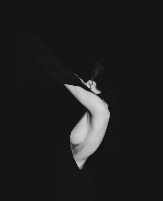 A woman's back and side are dramatically lit against a dark background, with a dark fabric obscuring part of her body, illustrating the theme of disappearing and finding oneself.