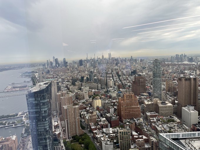 An aerial cityscape view of New York City taken from a high-rise window, showing Manhattan's skyline, the Hudson River, and reflections of contrails in the glass.
