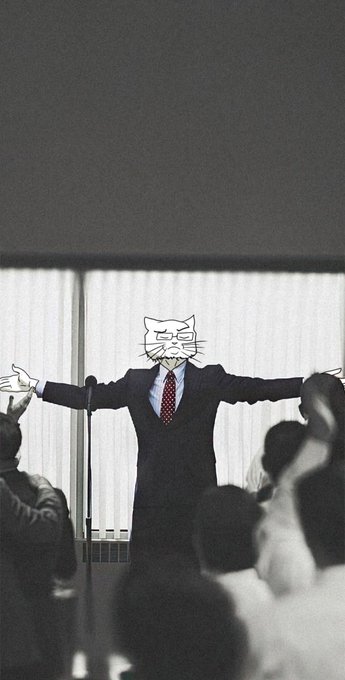 A figure in a suit standing with arms outstretched on a stage, with their face obscured by a cat emoji overlay.