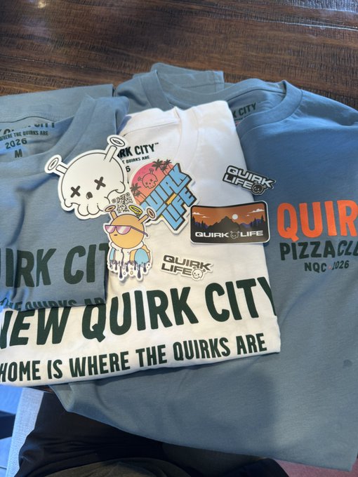 A pile of clothing merchandise featuring 'Quirk City' and 'Quirk Life' branding, including stickers with cartoon characters, patches, and t-shirts reading 'NEW QUIRK CITY' and 'HOME IS WHERE THE QUIRKS ARE'.