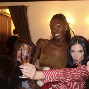 A smiling woman in an olive top toasts with a champagne glass between two friends, one wearing a red-and-white striped shirt, in a dimly lit indoor setting. The caption plays on the celebratory moment, humorously suggesting the woman once was hawking crypto but has clearly moved on to a more glamorous scene.