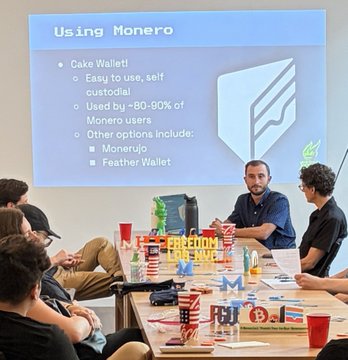 A meetup scene showing a presentation slide titled 'Using Monero' listing Cake Wallet as easy/self-custodial and used by ~80-90% of Monero users, with attendees seated around a table with a 'FREEDOM FOR ALL' sign.