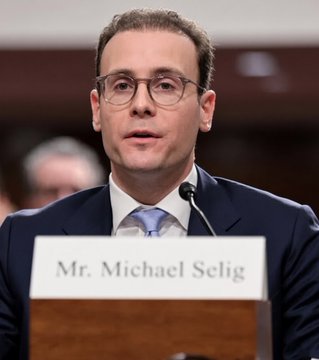 A photograph of a man wearing glasses and a dark suit speaking at a podium, with a nameplate reading 'Mr. Michael Selig'.