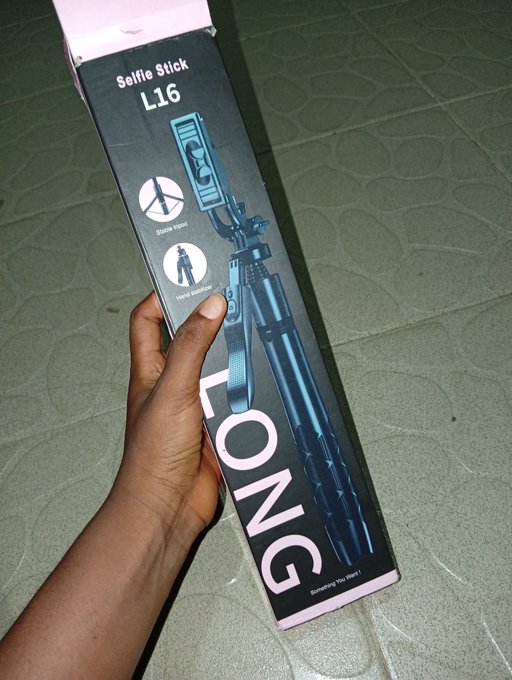 A hand holds a black product box for a 'Selfie Stick L16' by the brand FONG, displayed outdoors on a tiled surface.