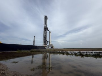A rocket on a launch pad stands against a cloudy sky, its reflection visible in a shallow pool of water in the foreground.