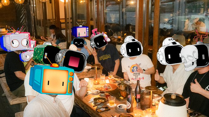 A group of people at a dinner table wearing various large, colorful robot/TV head masks, giving thumbs up while surrounded by food and drinks.