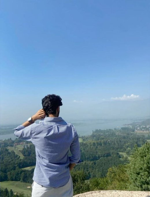 A man in a light blue striped shirt stands with his back to the camera, gazing out over a scenic landscape of lush green hills, a winding river, and a bright blue sky. The view complements the "Good morning" post, evoking a fresh, optimistic start to the day while tagging @NomismaNetwork × @useTria.