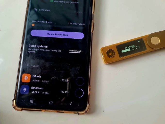 A smartphone displays the Ledger app interface showing 2 app updates in progress for Bitcoin (v2.5.0) and Ethereum (v1.22.3), alongside a gold Ledger hardware wallet showing "Processing" on its screen. This visually supports the message about updating Ledger wallets to patch security vulnerabilities.