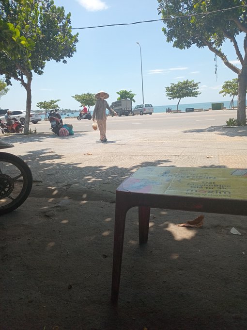 A street scene showing a person wearing a conical hat walking along a sunny coastal road, with a colorful table in the foreground and trees, motorcycles, and the ocean visible in the background.