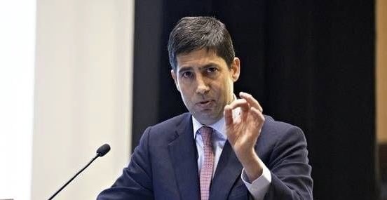 A man in a dark suit and pink tie speaks at a podium, gesturing with his right hand raised in an 'OK' sign.