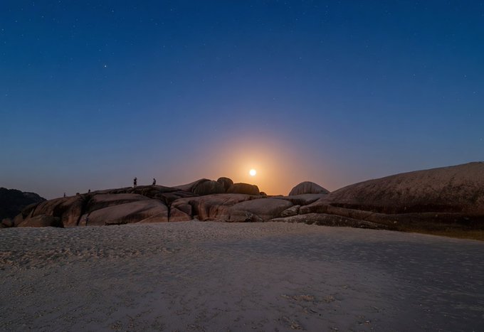 A serene twilight landscape photograph showing a full moon rising over a rocky, granite terrain with a sandy beach in the foreground and a few small figures standing on the boulders under a starry sky.