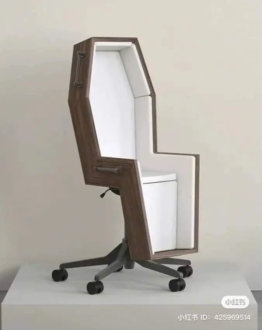 A coffin-shaped office chair with white cushioned interior and wooden exterior, mounted on a standard black wheeled office base, displayed in a gallery setting.