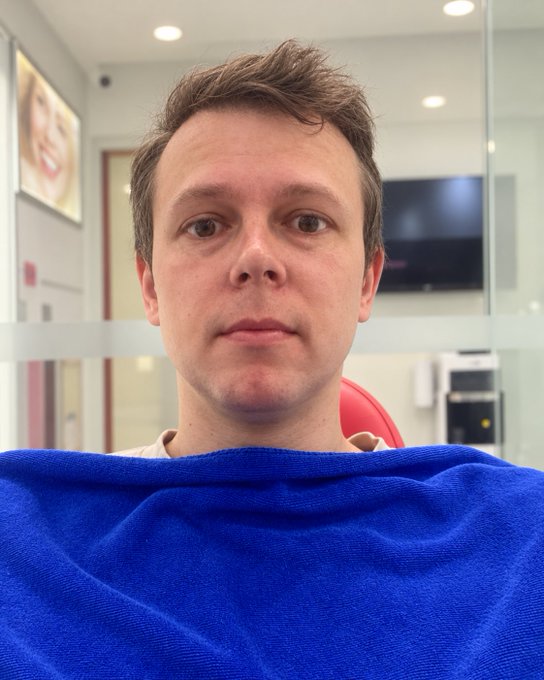 A selfie of a young man in a dental or medical office chair wearing a blue patient drape.