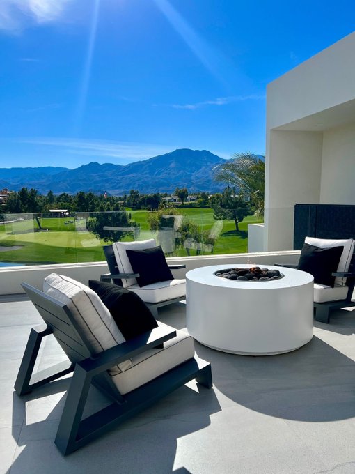 The image shows a patio with three chairs and a white circular table placed outside. The chairs are black and white striped, adding a stylish touch to the outdoor space. A fire pit is also present on the patio, providing warmth during colder evenings. The patio overlooks a golf course, offering an excellent view for relaxation and enjoyment of nature.