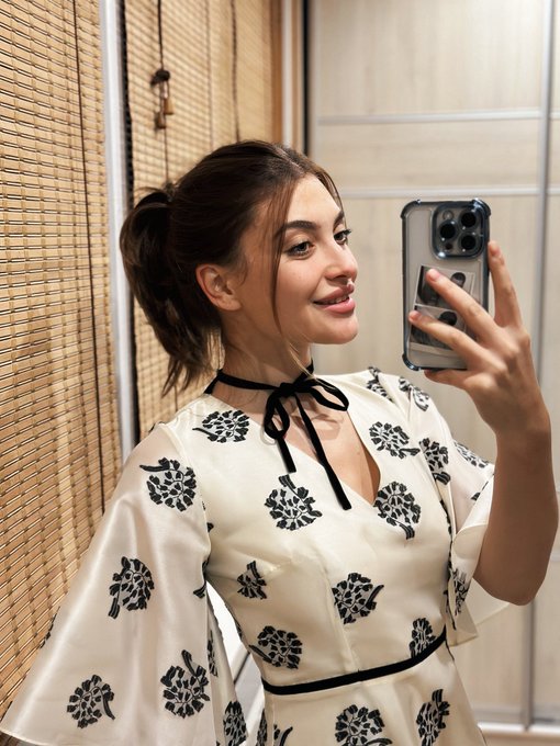 A woman with dark hair in a ponytail takes a selfie in a mirror, wearing a white dress with black floral patterns and a black ribbon choker.