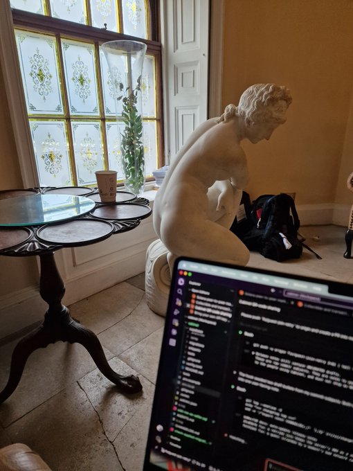 A photograph of a classic white marble statue of a woman bathing, located in a room next to a stained-glass window and a dark wooden table, with a laptop displaying code in the foreground.