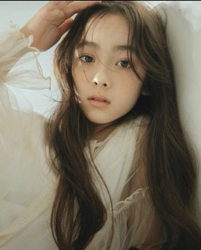 A close-up portrait of a young woman with long brown hair wearing a sheer white blouse and looking softly at the camera.