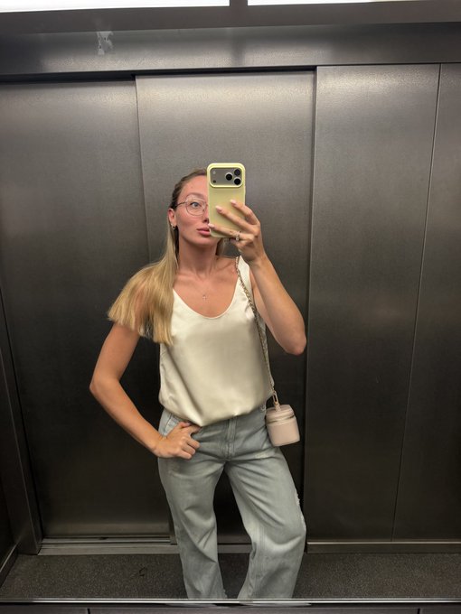 A mirror selfie shows a blonde woman wearing glasses, a white satin camisole, and light blue jeans, carrying a small round white handbag, standing in an elevator.