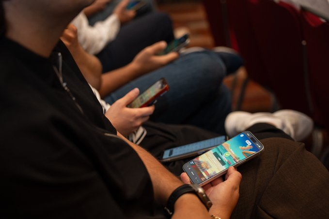 A group of community members are seen using their smartphones, including what appears to be the KuCoin app, at a gathering. The scene captures attendees engaging with crypto-related content and games during the meetup.
