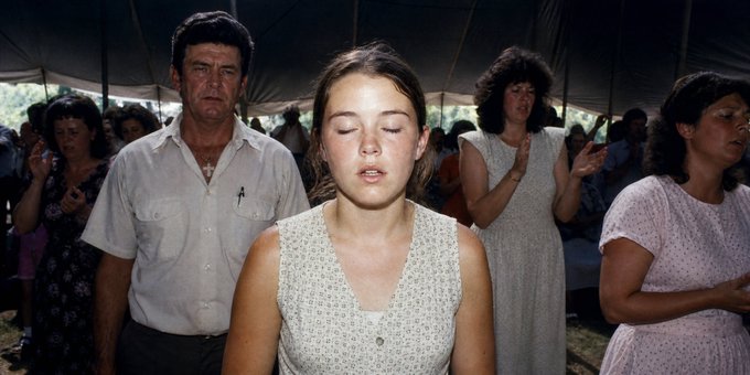 A group of people are praying under a tent, with a young woman in the center with her eyes closed and a man in a white shirt standing behind her, reminiscent of a scene from a film.